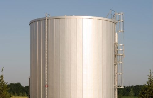 A tall, ribbed metal water storage tank with a caged ladder, standing in a grassy field flanked by evergreen trees.