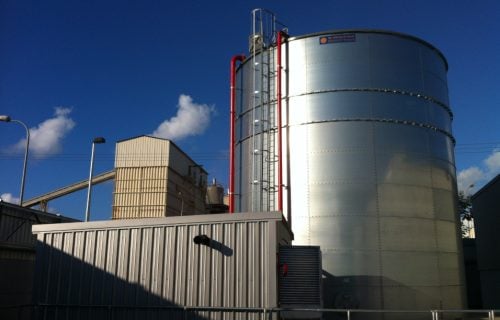 A galvanized steel water storage tank with red pipes and a ladder, set against a deep blue sky at an industrial facility.
